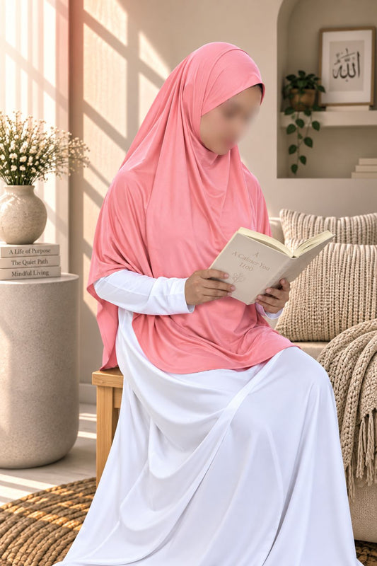 A woman wearing a pink hijab and a white long-sleeve dress sits on a wooden chair in a sunlit living room, holding an open book. The background includes a beige sofa, a plant, and a decorative side table.