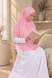 A woman wearing a light pink hijab and a white maxi dress sits on a beige sofa in a bright, modern living room, holding an open book.